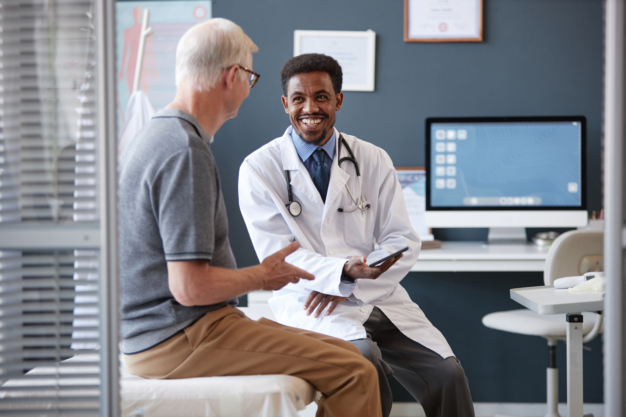 Family Practice - Smiling Black Doctor Holding Tablet Talking to Senior Patient in Consultation