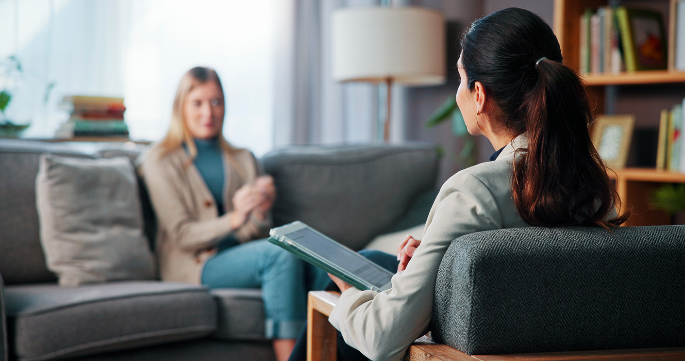 Consultation, therapy and woman with anxiety, stress and tablet for recovery and mental health in clinic. Counselor, psychologist and patient on sofa for trauma, diagnosis or conversation for help