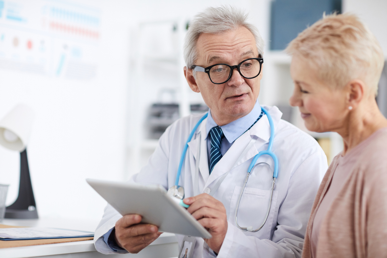 Oncology - Serious wrinkled senior doctor in lab coat sitting at table and using digital tablet with online patients records while giving recommendations to mature woman
